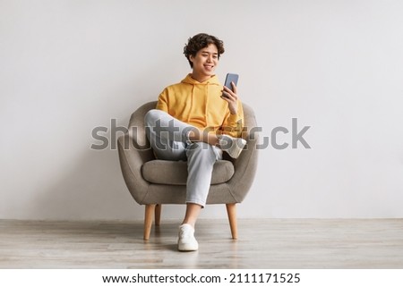 Handsome young Asian guy using cellphone, surfing web or social media, sitting in armchair, enjoying contemporary technologies, checking new mobile app against white studio wall