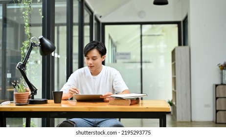 Handsome Young Asian Male Freelance Graphic Designer Or Office Worker In Casual Outfit Using Digital Tablet At His Office Desk.