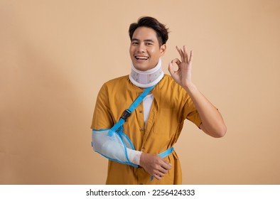 Handsome young asian man with broken arm in soft splint suffering a sore arm showing ok sign isolated on beige background, accident insurance concept. - Shutterstock ID 2256424333