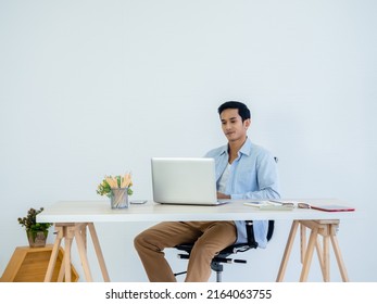 Handsome young Asian man in denim shirt using laptop on table on white clean space wall background in home office. Learning, working, startup with small business, stock, financial, investment concept. - Powered by Shutterstock