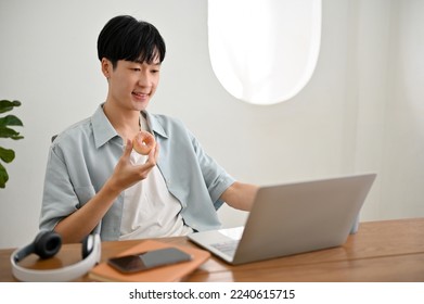 Handsome young Asian man enjoys eating donut while working on his project on his laptop computer. - Powered by Shutterstock