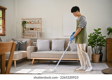 Un hombre apuesto y joven asiático que se centra en limpiar su sala de estar, usando una aspiradora para limpiar la alfombra. día de limpieza, tareas domésticas, concepto de limpieza