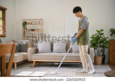 Handsome young Asian man focusing on cleaning his living room, using stick vacuum cleaner to clean the carpet. cleaning day, house chores, housekeeping concept