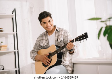 handsome, young asian man looking away while playing acoustic guitar at home