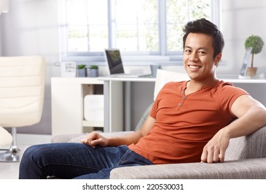 Handsome Young Asian Man Sitting At Home On Sofa, Smiling Happy, Looking At Camera.