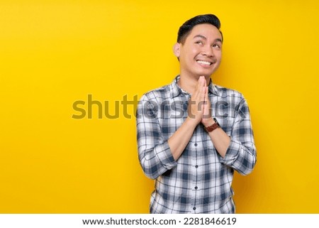 Handsome young Asian man trustfully pleaded in plaid shirt while hands folded in a prayer gesture, begging about something isolated on yellow background. people lifestyle concept