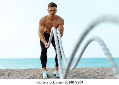 Handsome young athlete man doing fitness workout, by the sea on the beach on a sunny day, using two fighting ropes on the beach, morning workout - Powered by Shutterstock