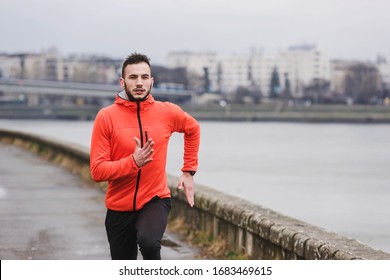 Handsome Young Athlete Man Running Fast Along River In Orange Windbreaker Jacket. Extreame Weather Sport. Running On A Rainy Day.