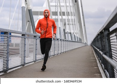 Handsome young athlete running fast along big modern bridge in orange windbreaker jacket. Exercising, Jogging, Sport, Winter. Male athlete running. - Powered by Shutterstock