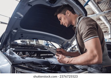 Handsome young auto mechanic in uniform is making notes while examining car in auto service - Powered by Shutterstock