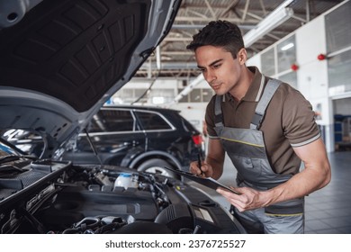 Handsome young auto mechanic in uniform is making notes while examining car in auto service - Powered by Shutterstock