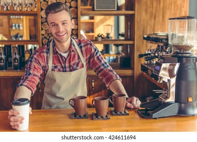 Handsome young barista in apron is giving a cup of coffee, looking at camera and smiling while standing at the bar counter at cafe