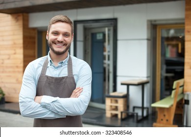 Handsome young barista is standing near his cafe outdoors. He is looking forward and smiling. The man crossed his arms with confidence