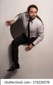 Handsome Young Bearded Businessman Climbing Out Of A Circular Hole In A White Wall In A Handsome Young Bearded Businessman Climbing Out Of A Circular Hole In A White Wall In A