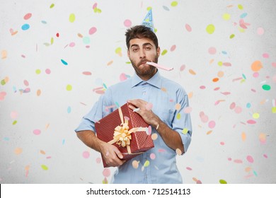 Handsome Young Bearded Caucasian Male In Formal Shirt Blowing Party Horn And Holding Present In Wrapping With Ribbon, Going To His Colleague To Celebrate Birthday, Having Confident Expression