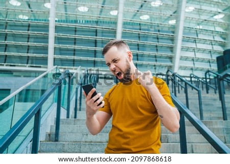 Handsome young bearded football fan celebratin money win after betting at favourite team using bookmaker's mobile application, stadium on the background.