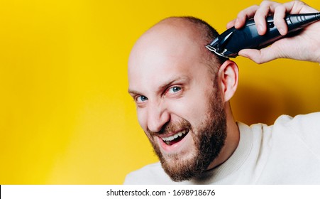 Handsome Young Bearded Man Emotional Trimming His Head With A Trimmer On Yellow Background. Shaving Head At Home In Quarantine. Selfisolated Man Is Shaving His Beard On Yellow Background.