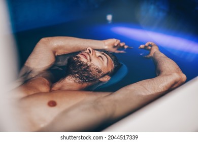 Handsome young bearded man floating in tank filled with dense salt water used in meditation, therapy, and alternative medicine. 