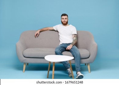 Handsome Young Bearded Man Guy In White Blank Empty T-shirt Sitting On Couch Isolated On Pastel Blue Wall Background Studio Portrait. Sport Leisure Lifestyle Concept. Cheer Up Support Favorite Team