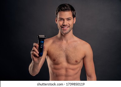 Handsome young bearded man isolated. Portrait of topless muscular man is standing on gray background with electric Shaver in hand. Men care concept
