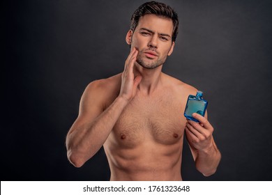 Handsome young bearded man isolated. Portrait of shirtless muscular man is standing on grey background and using face aftershave lotion. Men care concept