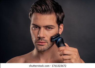 Handsome young bearded man isolated. Portrait of topless muscular man is standing on gray background with electric Shaver in hand while shaving. Men care concept