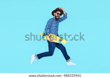 Handsome young bearded man jumping with yellow skateboard against the colorful wall. Hipster with beard in motion on blue background