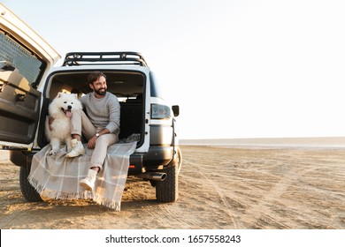 Handsome Young Bearded Man Sitting In The Back Of His Car, Playing With Dog At The Beach