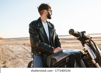 Handsome young bearded man wearing leather jacket sitting on a motorbike at the beach
