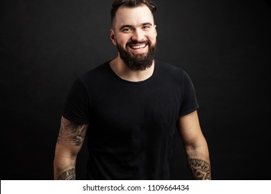 Handsome young biker with tattooed hands in casual black wear smiling while standing against black background. Positive Emotions. Sthength, Power, Humanity and People Concept. - Shutterstock ID 1109664434
