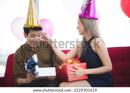Handsome young birthday man wearing cap holding and opening gift while sitting besides beautiful girlfriend holding gift and teasing him by pulling his ears and sitting on couch at home party