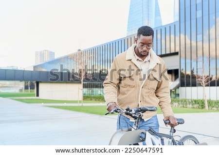Handsome young black college student parking his bicycle. He is wearing blue jeans and a jacket. Bike as conveyance at the university campus.