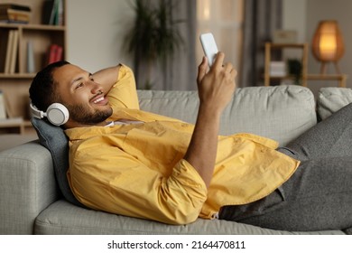 Handsome Young Black Guy In Wireless Headphones Listening To Music Online On Smartphone, Lying On Couch At Home. Happy African American Man In Headset Watching Videos On Mobile Phone