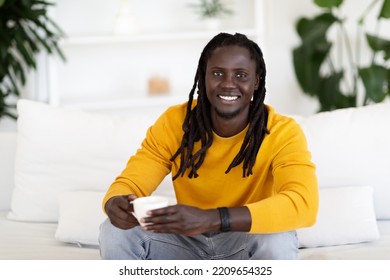 Handsome Young Black Man Drinking Coffee While Relaxing On Couch At Home, Smiling African American Man With Dreadlocks Posing With Cup Of Tea In Hands In Cozy Living Room Interior, Copy Space