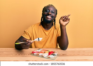 Handsome Young Black Man Eating Sushi Sitting On The Table Smiling Happy Pointing With Hand And Finger To The Side 