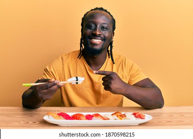 Handsome Young Black Man Eating Sushi Sitting On The Table Smiling Happy Pointing With Hand And Finger 