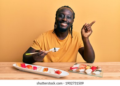 Handsome Young Black Man Eating Sushi Sitting On The Table Smiling Happy Pointing With Hand And Finger To The Side 