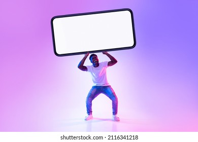 Handsome Young Black Man Holding Huge Heavy Smartphone With Empty White Screen In Neon Light, Mockup For Advertisement, Mobile App Or Website Design. Cellphone Display Template