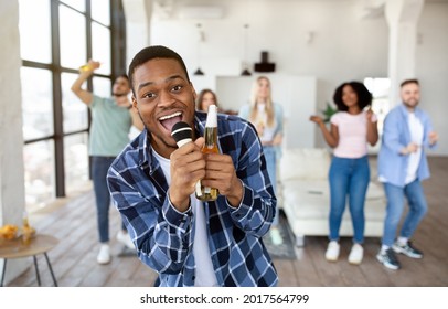 Handsome Young Black Man Singing Karaoke At Home, Holding Bottle Of Beer And Mic, Dancing And Having Fun With Group Of Friends. Cheerful Millennial Guy Partying With Fellow Students Indoors