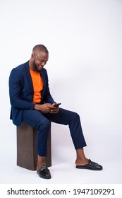 Handsome Young Black Man Wearing A Suit Using His Phone, Sitting Against A White Plain Background