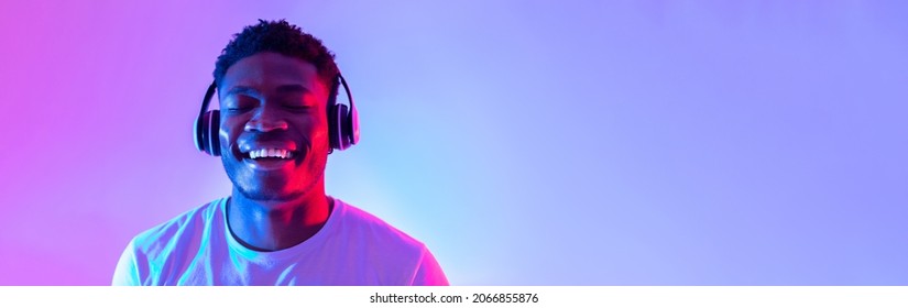 Handsome young black man wearing headphones, listening to music with closed eyes in neon light, banner design. Happy African American guy enjoying new playlist and smiling