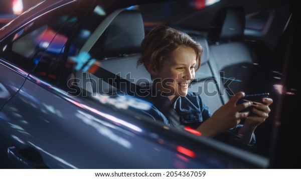 Handsome Young Boy is\
Sitting on Backseat of a Car, Commuting Home at Night. Passenger\
Playing Video Game on Smartphone while in Taxi in City Street with\
Working Neon Signs.