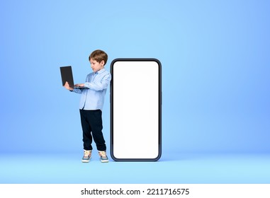 Handsome Young Boy Wearing Formal Wear Is Standing Holding Laptop Near Large Empty White Mockup Smartphone Case In Background. Blue Wall. Concept Of Gadgets, Learning, Studying, Progressive Kids