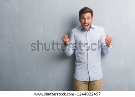 Handsome young business man over grey grunge wall wearing elegant shirt celebrating surprised and amazed for success with arms raised and open eyes. Winner concept.