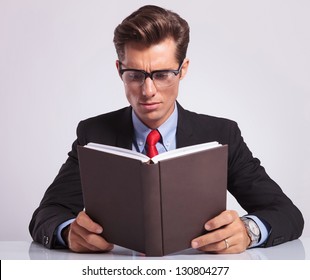 Handsome Young Business Man Reading A Book At His Desk, On Gray Background