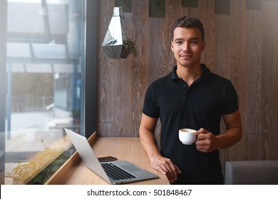 Handsome young business man standing with laptop and looking to camera while holding a cup of coffee