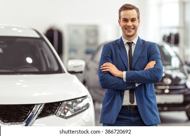 Handsome young businessman in classic blue suit is smiling and looking at camera while standing near the car in a motor show