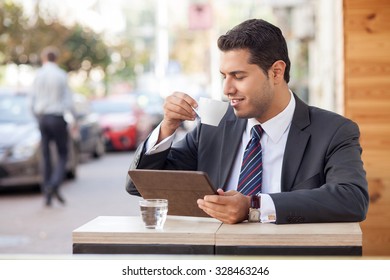 Handsome young businessman is drinking coffee in cafeteria outdoors. His eyes are closed with pleasure. He is sitting at table and using tablet. The man is smiling - Powered by Shutterstock