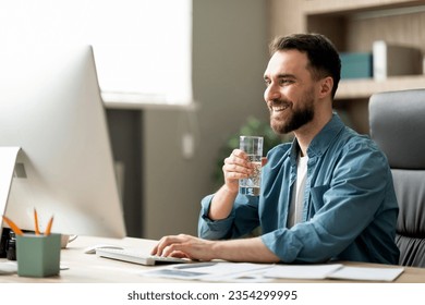 Handsome young businessman drinking water and working on computer in office, smiling millennial male entrepreneur enjoying refreshing drink at workplace and looking at pc monitor, copy space - Powered by Shutterstock