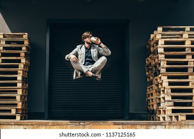 handsome young businessman floating in air in lotus pose and drinking coffee at industrial location in front of roller gate 